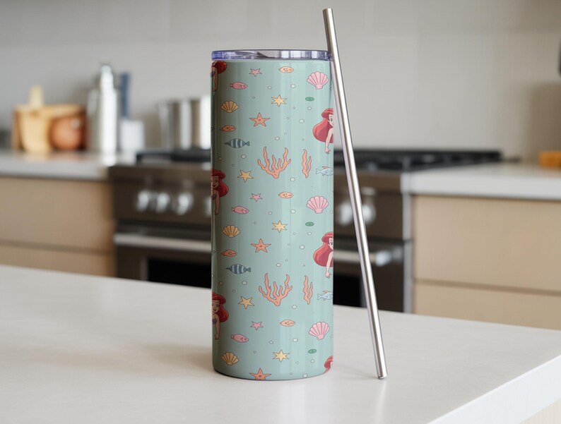 May include: A tall, cylindrical tumbler with a light teal background featuring a repeating pattern of a mermaid, seashells, starfish, coral, and fish. A silver-colored straw rests against the side of the tumbler. The tumbler is designed for cold beverages.