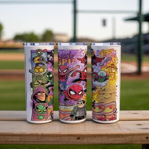 May include: Three stainless steel tumblers with colourful Spider-Man comic book art. Each tumbler features a different design with various characters and action scenes. The tumblers are cylindrical with silver lids, set on a wooden surface.
