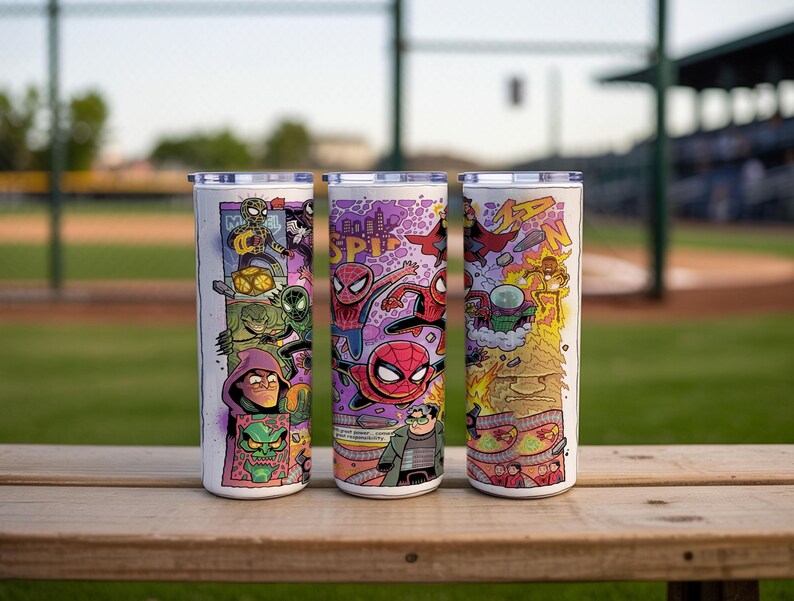 May include: Three stainless steel tumblers with colourful Spider-Man comic book art. Each tumbler features a different design with various characters and action scenes. The tumblers are cylindrical with silver lids, set on a wooden surface.