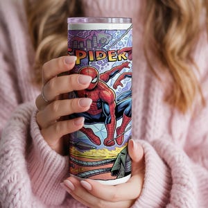 May include: A cylindrical tumbler featuring a vibrant comic book graphic of Spider-Man in a red and blue suit. The design includes a city skyline, the partial text SPIDER, and mechanical tentacles at the bottom against a purple and blue background.