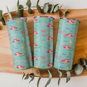 May include: Three teal insulated tumblers with a mermaid pattern. The design features red-haired mermaids, pink starfish, yellow seashells, and green seaweed. The tumblers are cylindrical and have silver lids.