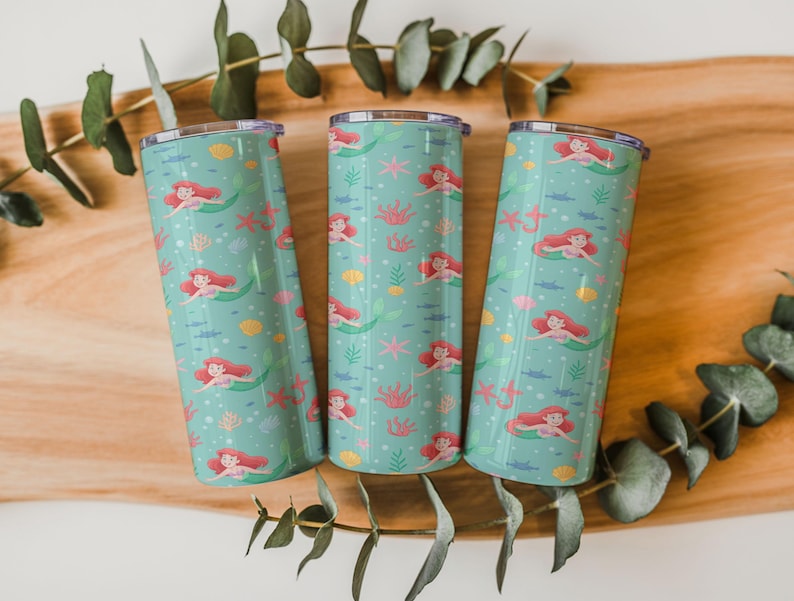 May include: Three teal insulated tumblers with a mermaid pattern. The design features red-haired mermaids, pink starfish, yellow seashells, and green seaweed. The tumblers are cylindrical and have silver lids.