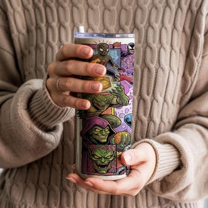 May include: A cylindrical stainless steel tumbler featuring a colorful comic book style graphic print of various characters including a spider-themed hero, a green reptilian creature, and a green-faced villain with a pointed purple hood.