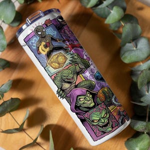 May include: A cylindrical white tumbler featuring a colorful graphic print of various comic book characters, including a masked hero in a black suit, a green reptilian creature, and a green-skinned villain wearing a purple hood and cape.