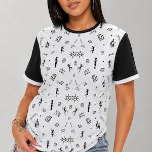 May include: A short-sleeved t-shirt with a white front panel featuring a black geometric and tribal-inspired pattern of figures and symbols. The garment has a solid black crew neckline, solid black sleeves, and white trim at the sleeve cuffs.