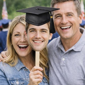 May include: A graduation-themed photo featuring a smiling man's face on a stick, topped with a black mortarboard and tassel. Two adults smile broadly, embracing the graduate. The background suggests an outdoor graduation ceremony.