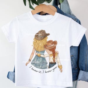 May include: A white short-sleeved t-shirt featuring a graphic illustration of two figures wearing cowboy hats with their arms around each other. The text below the figures reads, I know it, I know you. The shirt is displayed on a wooden hanger.