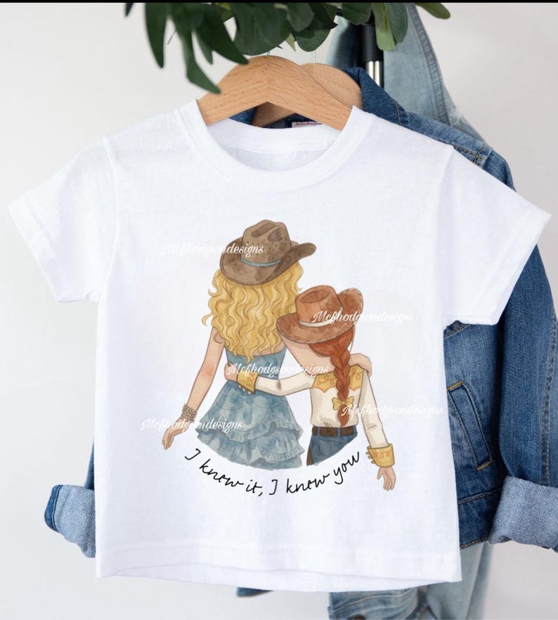 May include: A white short-sleeved t-shirt featuring a graphic illustration of two figures wearing cowboy hats with their arms around each other. The text below the figures reads, I know it, I know you. The shirt is displayed on a wooden hanger.