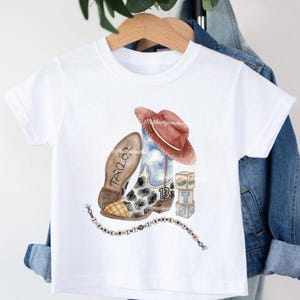 May include: A white short-sleeve t-shirt featuring a watercolor graphic of a cowboy boot with a cow print, a brown hat, the number 13, blocks with the letter T, and a string of beads with the text I KNEW YOU WERE TROUBLE and TAYLOR on the sole.