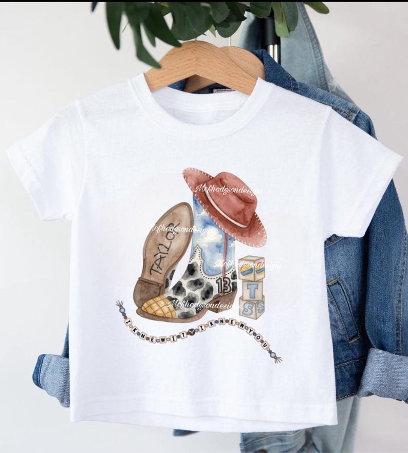 May include: A white short-sleeve t-shirt featuring a watercolor graphic of a cowboy boot with a cow print, a brown hat, the number 13, blocks with the letter T, and a string of beads with the text I KNEW YOU WERE TROUBLE and TAYLOR on the sole.