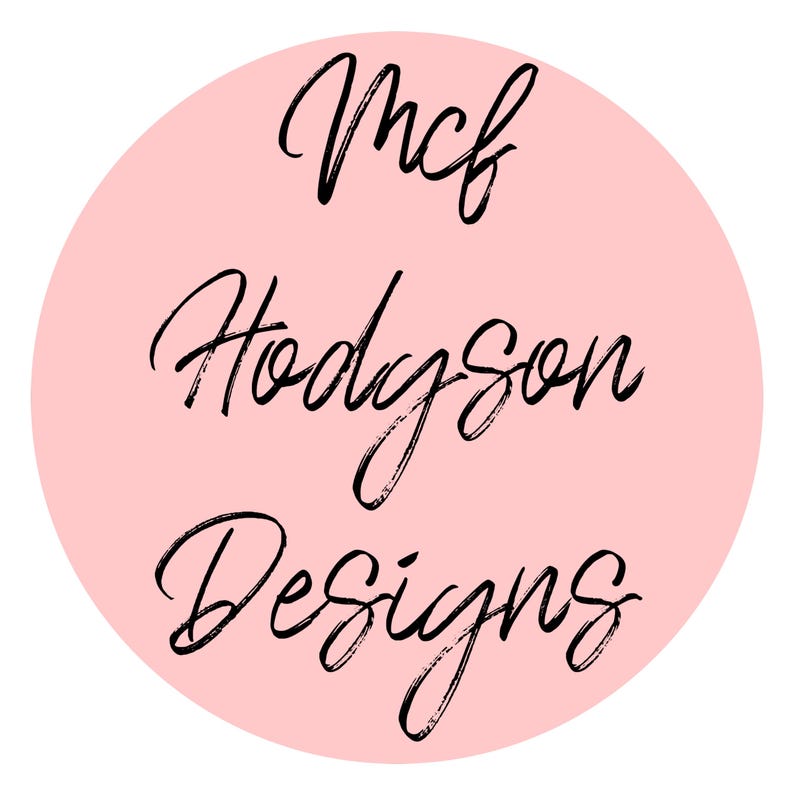 May include: A round, light pink logo with the words "Mcf Hodgson Designs" in black, handwritten-style script. The text is arranged in three lines, centered within the pink circle, against a white background.