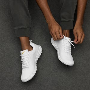 May include: White trainers with a textured design and white laces. The trainers have a yellow accent on the side. The person is tying the laces of the trainers. The trainers are on a grey surface.