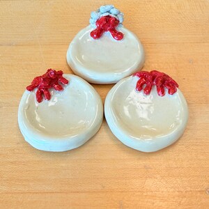 Puede incluir: Tres platos de cerámica hechos a mano. Los platos son de color blanquecino con un diseño rojo texturizado en la parte superior. El plato superior tiene un acento azul. Los platos están dispuestos sobre una superficie de madera marrón claro.