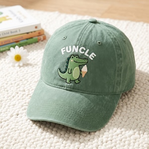 May include: A sage green baseball cap featuring an embroidered graphic of a light green crocodile holding a tan ice cream cone. Above the crocodile, the word "FUNCLE" is embroidered in white capital letters. The cap has a curved brim.