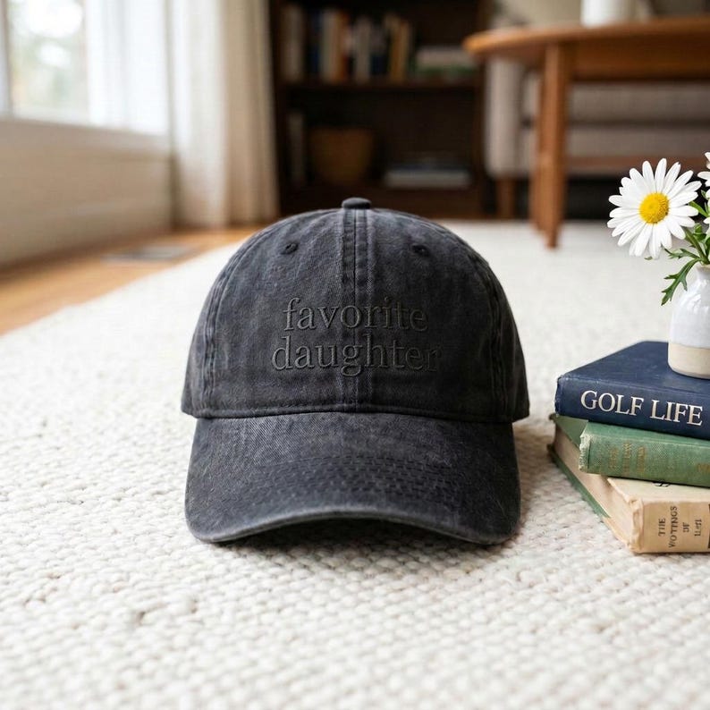May include: A black, distressed baseball cap with the embroidered text "favorite daughter". The hat is sitting on a white rug. The hat is a classic six-panel design with a curved brim.