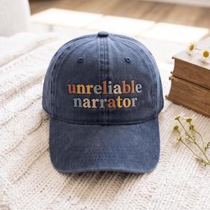 Puede incluir: Una gorra de béisbol azul marino con construcción de seis paneles y visera curva. La parte frontal presenta el texto bordado "unreliable narrator" en letras minúsculas alternadas en naranja, azul claro y crema en el centro.