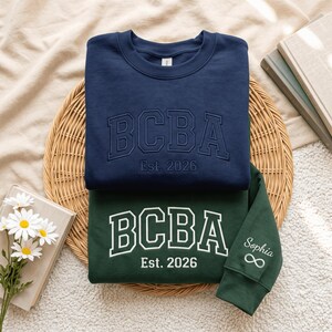 Könnte beinhalten: Ein marineblaues Sweatshirt mit tonaler Stickerei BCBA Est. 2026 und ein dunkelgrünes Sweatshirt mit weißer Stickerei BCBA Est. 2026, wobei der Name Sophia und ein Unendlichkeitssymbol auf dem Ärmelbündchen eingestickt sind.