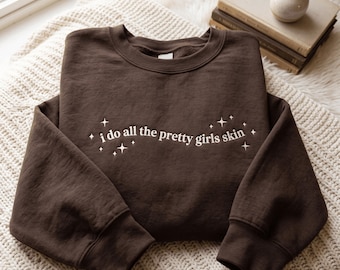 Puede incluir: Una sudadera de cuello redondo en color marrón oscuro con el texto bordado en blanco "i do all the pretty girls skin" centrado en el pecho, rodeado de pequeños iconos de estrellas bordados dispuestos en un patrón ondulado en la parte frontal.