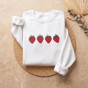May include: A white long-sleeve crewneck sweatshirt featuring an embroidered design of four red strawberries with green leaves arranged in a horizontal row across the center of the chest. The garment has ribbed cuffs and a ribbed neckline.