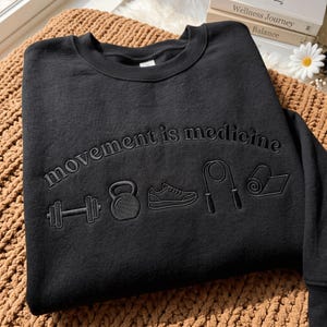 以下が含まれることがあります： 「movement is medicine」という刺繍文字が曲線状に施された黒いクルーネックのスウェットシャツ。文字の下には、ダンベル、ケトルベル、スニーカー、縄跳び、丸めたヨガマットの刺繍アイコンが黒い糸でデザインされています。