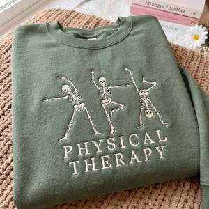 May include: An olive green crewneck sweatshirt featuring an embroidered design of three white skeletons in various yoga poses. Below the skeletons, the text "PHYSICAL THERAPY" is embroidered in a clean, white, sans-serif font on the chest.