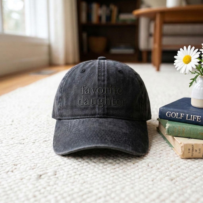 May include: A black, distressed baseball cap with the embroidered text "favorite daughter". The hat is sitting on a white rug. The hat is a classic six-panel design with a curved brim.