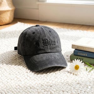 May include: A dark gray baseball cap with the embroidered text "I Bite" in a gothic font. The cap is sitting on a white rug next to a stack of books and a daisy.