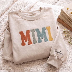 以下が含まれることがあります： ベージュ色のクルーネックのスウェットシャツ。胸元には「MIMI」という文字が、錆色、セージグリーン、青、マスタードイエローの異なる色で大きく刺繍されています。袖には黒い糸で「Sophia」という名前が刺繍されています。