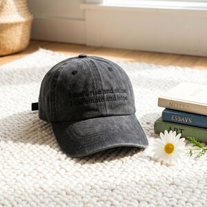 May include: A charcoal gray baseball cap with the embroidered text "I don't rise and shine. I caffeinate and hope." The cap is sitting on a white rug next to a stack of books and a daisy.