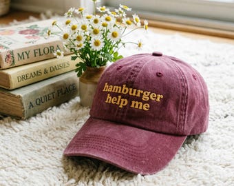 May include: A burgundy baseball cap with the embroidered text "hamburger help me" in yellow. The cap is sitting on a white shag rug, near a vase of daisies and a stack of vintage books.