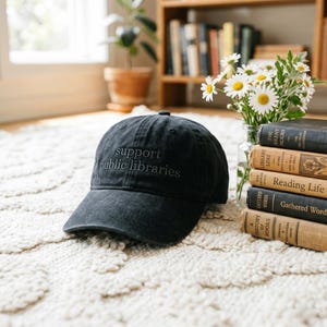 May include: A black baseball cap with the text support public libraries embroidered in a matching dark thread on the front panel. The cap features a structured crown and a curved brim, presented alongside a stack of vintage books and flowers.