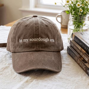 Può includere: Un cappellino da baseball marrone con il testo ricamato in bianco "in my sourdough era" centrato sul pannello frontale. Il cappellino presenta una corona strutturata, una visiera curva e cuciture visibili sui pannelli e sulla visiera.