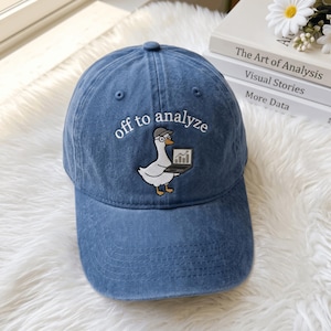 May include: A blue denim baseball cap featuring an embroidered graphic of a white goose wearing a grey cap and glasses while holding a laptop. The text "off to analyze" is embroidered in white above the goose on the front of the cap.