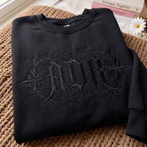 Puede incluir: Una sudadera negra de cuello redondo con bordado negro tonal en el pecho. El diseño consiste en las letras ADR en una fuente gótica estilizada, rodeadas de intrincados patrones de enredaderas espinosas que se extienden desde las letras.