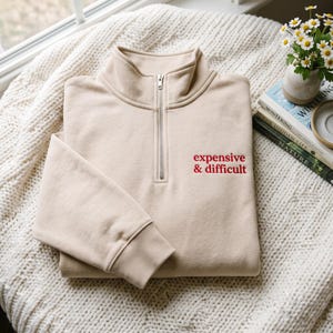 Può includere: Una felpa beige con zip a un quarto, collo alto e cerniera in metallo argentato. Il testo "expensive & difficult" è ricamato con filo rosso sulla parte superiore destra del petto. Il capo presenta maniche lunghe con polsini a costine.