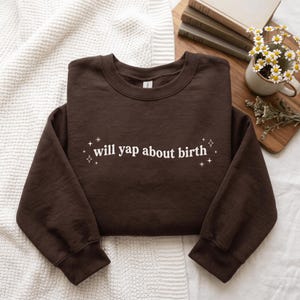 May include: A dark brown crewneck sweatshirt featuring the white text will yap about birth centered on the chest, accented by small white star-shaped graphics on either side of the lettering. The garment has long sleeves and ribbed cuffs.