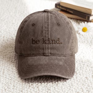Può includere: Un cappellino da baseball marrone scuro con una finitura consumata e slavata. La parte anteriore del cappellino presenta il testo ricamato "be kind." in filo marrone scuro. Il cappellino ha una corona strutturata, una visiera curva e un bottone.