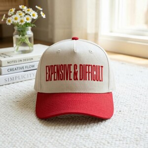 Peut inclure: Casquette de baseball beige avec une visière rouge et un bouton rouge. Le texte "EXPENSIVE & DIFFICULT" est brodé en rouge sur le devant. Le chapeau est posé sur un tapis blanc.