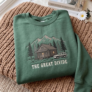 May include: A forest green crewneck sweatshirt featuring an embroidered graphic of a log cabin, pine trees, mountains, and an SUV. The text THE GREAT DIVIDE is embroidered in white below the cabin. The design includes shades of brown, grey, and white.