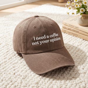 May include: A brown baseball cap featuring the embroidered white text "I need a coffee not your opinion" on the front panel. The cap has a curved brim, a button on the top, and stitched eyelets for ventilation, constructed from a solid fabric.