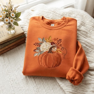 May include: A burnt orange crewneck sweatshirt featuring an embroidered design of a pumpkin topped with a bouquet of cream-coloured flowers, autumn leaves, and berries. The embroidery is centred on the chest of the long-sleeved garment.