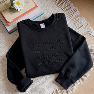 May include: A black crewneck sweatshirt featuring the embossed text "i survived my wife's doctorate program" in a matching black tone. The garment has long sleeves with ribbed cuffs and a ribbed neckline, presented in a clean, simple design.