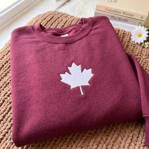 May include: A maroon crewneck sweatshirt featuring a white embroidered maple leaf centred on the chest. The garment has a ribbed collar, long sleeves, and a soft fabric texture. The design is simple, focusing on the contrast between the dark red fabric and the bright white stitching of the leaf emblem.
