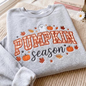 May include: A light gray crewneck sweatshirt featuring embroidered text that reads PUMPKIN season. The word PUMPKIN is in orange and white gingham block letters, while season is in black script. Small embroidered pumpkins, leaves, and a ghost.