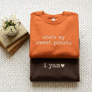 Könnte beinhalten: Ein orangefarbenes Sweatshirt mit Rundhalsausschnitt und dem weiß gestickten Text 'she's my sweet potato' sowie ein dunkelbraunes Sweatshirt mit Rundhalsausschnitt und dem weiß gestickten Text 'i yam' und einem kleinen weißen Herz.
