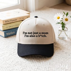 May include: A beige baseball cap with a black brim featuring the embroidered black text: i'm not just a mom i'm also a b*tch. The cap has a structured crown with a black button at the top center and stitched eyelets on the front panels.