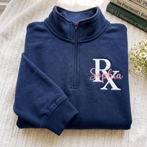 Può includere: Una felpa blu navy con zip a un quarto e un simbolo Rx bianco ricamato sul petto sinistro. Il nome Sophia è cucito in corsivo rosa chiaro al centro del simbolo Rx bianco. Il capo presenta un colletto alto e polsini a coste.