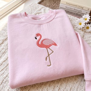Könnte beinhalten: Ein hellrosa Sweatshirt mit Rundhalsausschnitt und einer mittigen Stickerei eines Flamingos. Der Flamingo ist in Korallen- und Rosatönen gehalten, hat einen schwarz gespitzten Schnabel und lange, dünne beigefarbene Beine und steht auf einem Bein.