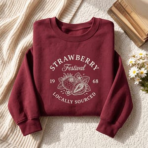 May include: A maroon crewneck sweatshirt featuring a white graphic print that reads Strawberry Festival 1968 Locally Sourced, accompanied by illustrations of whole and halved strawberries with leaves and small blossoms centered on the chest.