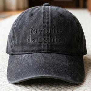 May include: A dark gray baseball cap with the embroidered text "favorite daughter" in a matching tone. The cap has a curved brim and a classic six-panel design, suitable for casual wear.